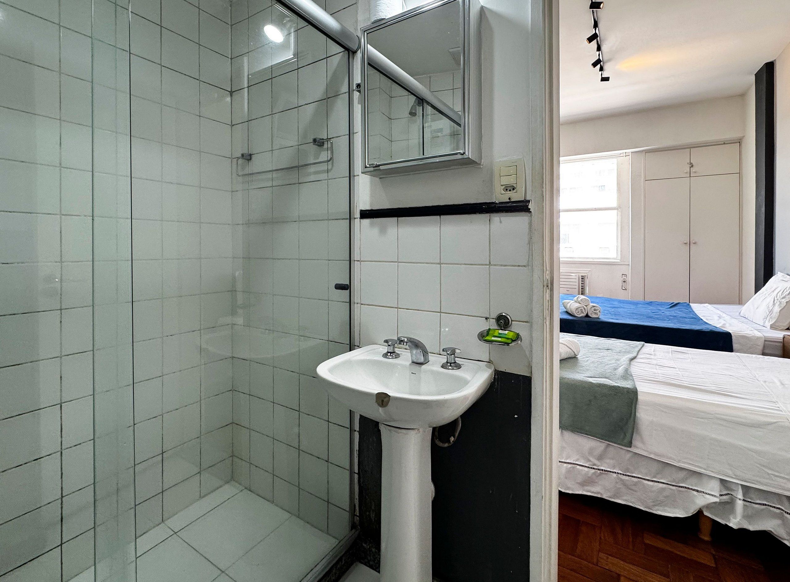 A bathroom with a white sink, a glass shower enclosure, and a bed with white bedding in the adjacent room.