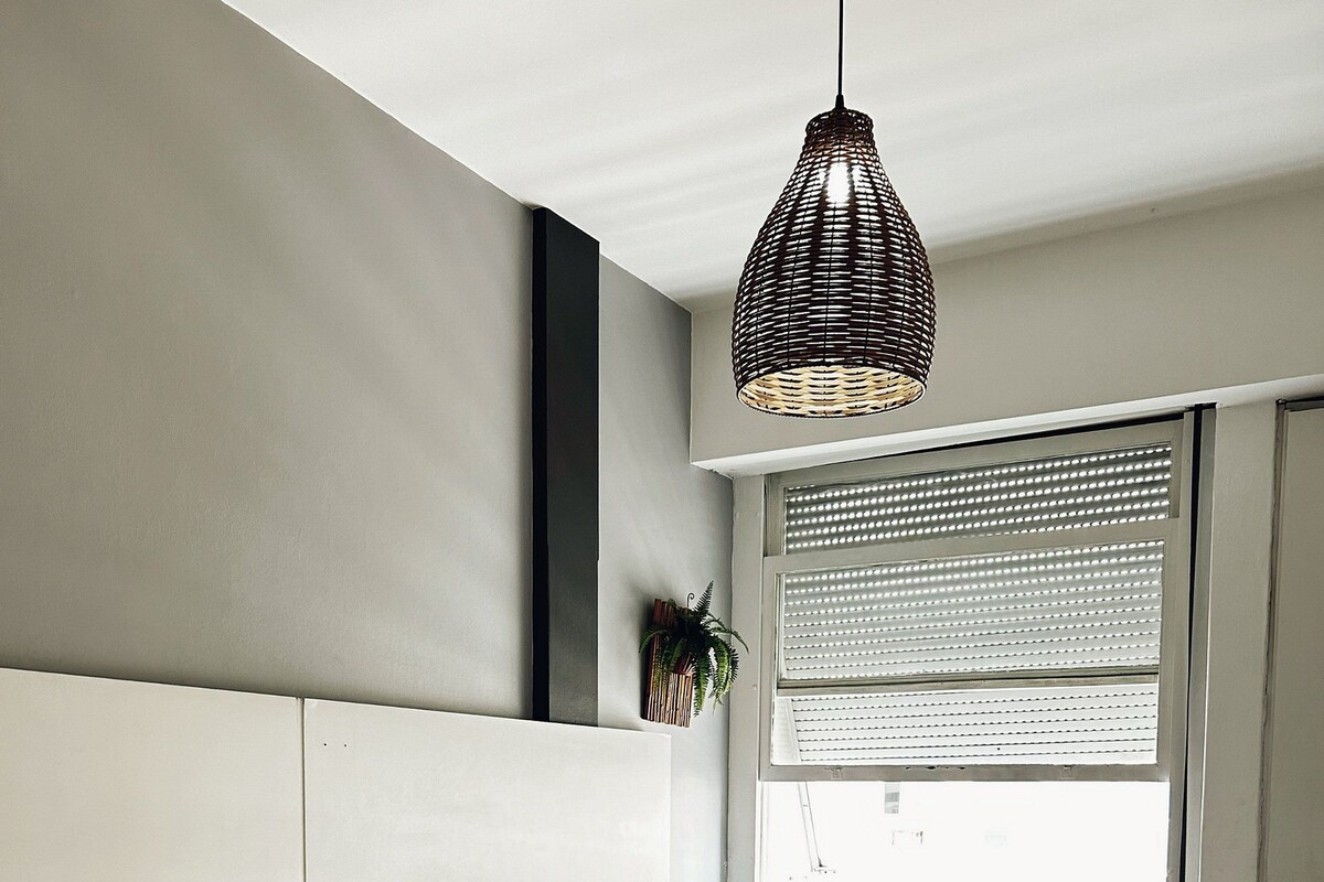 Une chambre avec deux lits, un luminaire suspendu, une fenêtre avec des stores et une plante accrochée au mur.