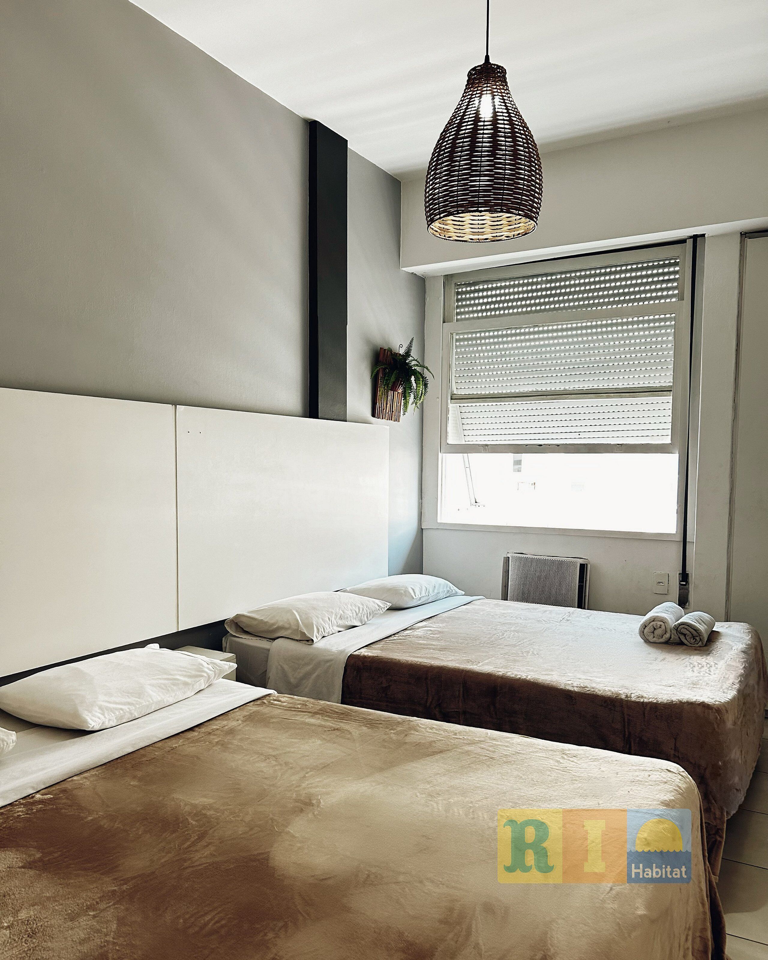 A bedroom with two beds, a hanging light fixture, a window with blinds, and a plant on the wall.