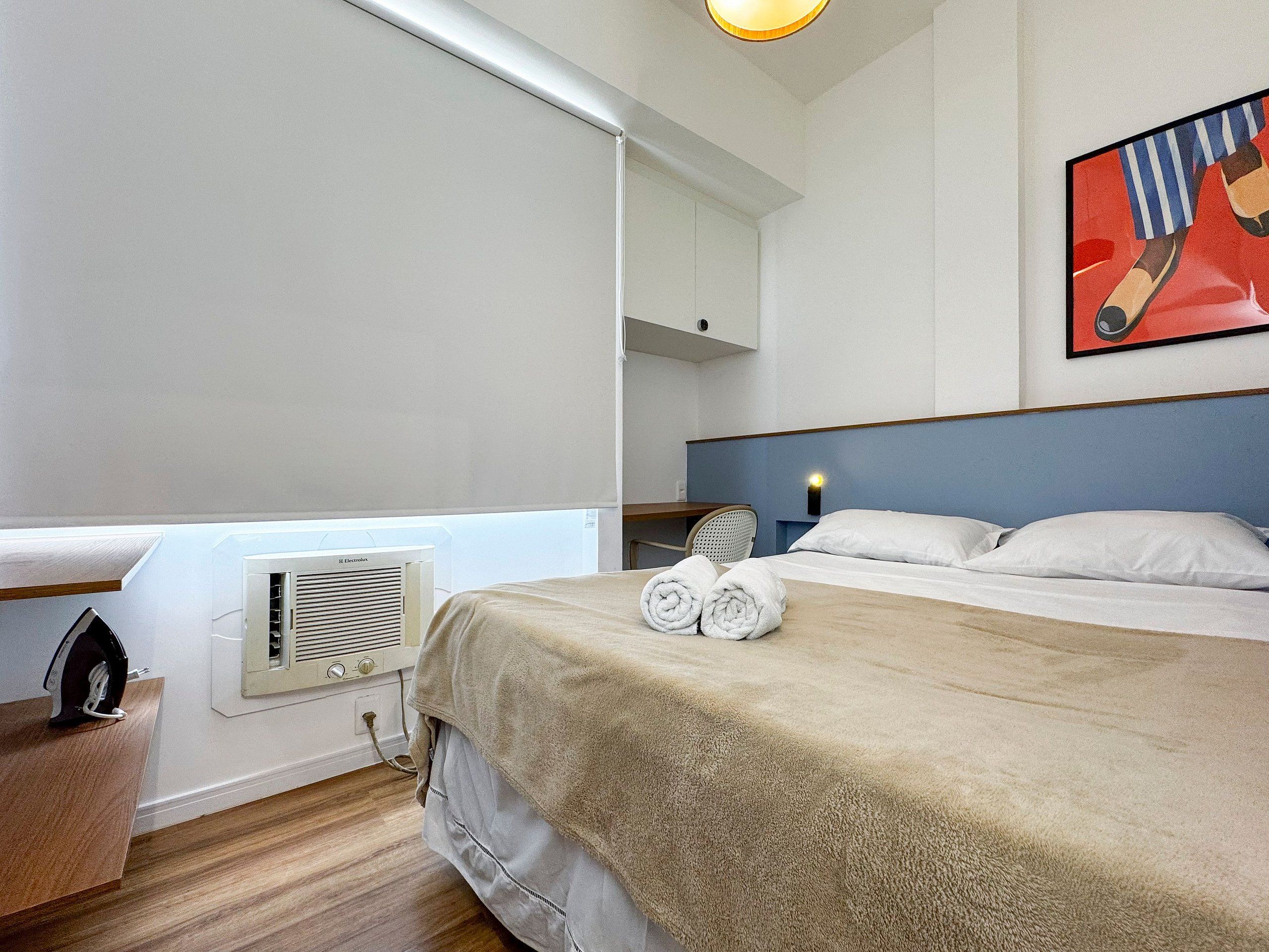 Habitación de hotel con cama, escritorio, silla, calefactor y un cuadro en la pared.