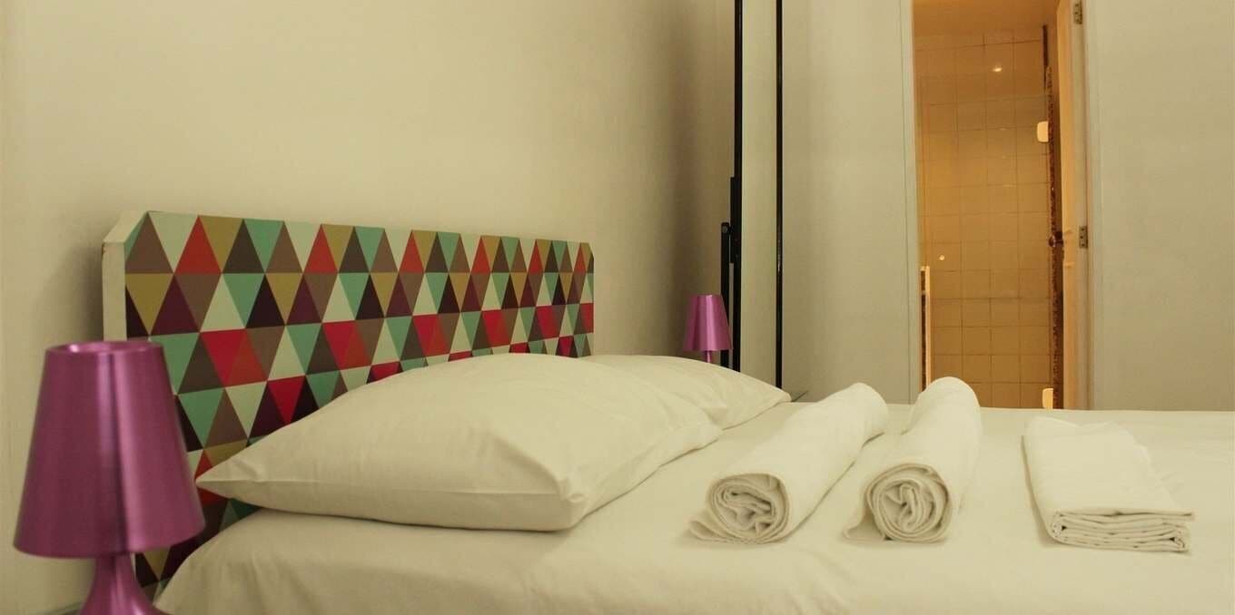 A hotel room with a bed, pillows, a headboard with a geometric pattern, a lamp, and a door.