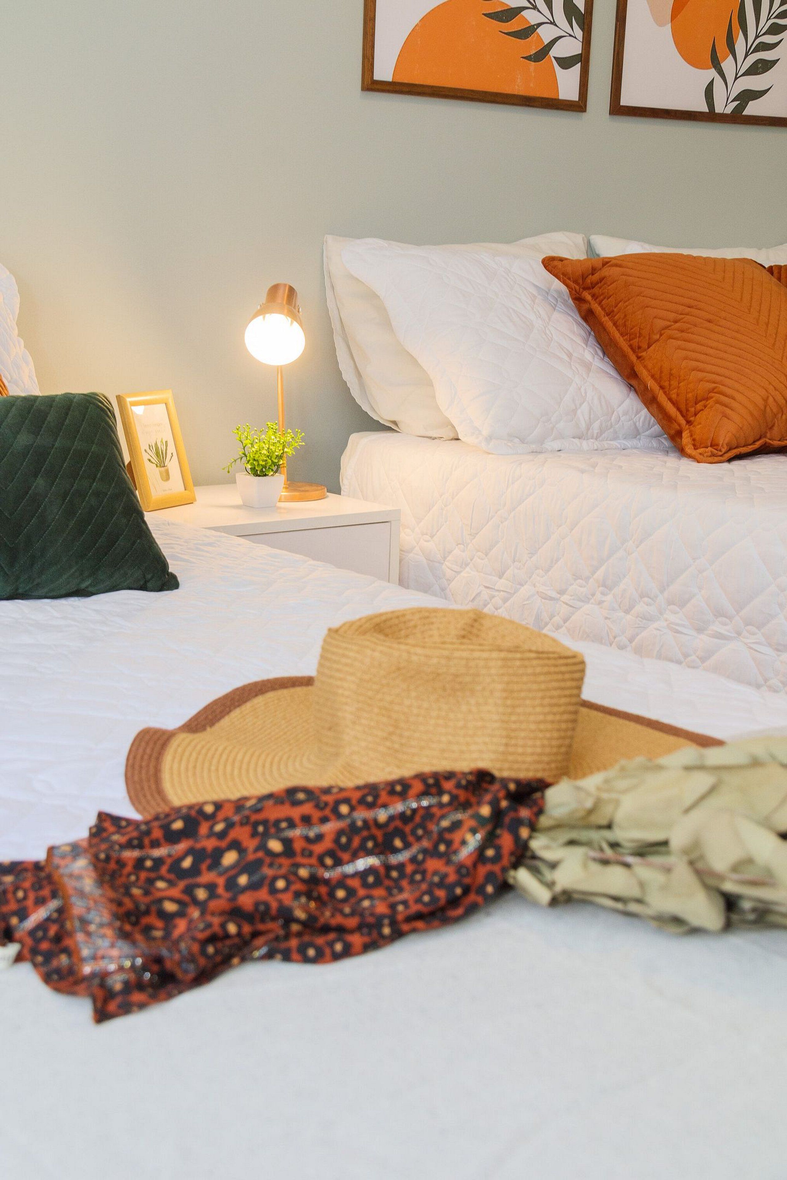 A neatly made bed with a white quilt, an orange pillow, and a green pillow. There is a bedside lamp, a framed picture, and a small potted plant on the nightstand.