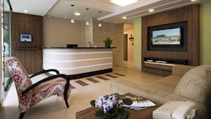 Zona de recepción de hotel con mostrador, un sofá, una silla, una mesa de centro con un jarrón de flores, un teléfono y un televisor montado en la pared.