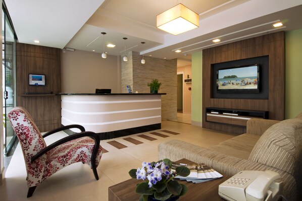 A hotel reception area with a reception desk, a sofa, a chair, a coffee table with a flower vase, a telephone, and a television mounted on the wall.