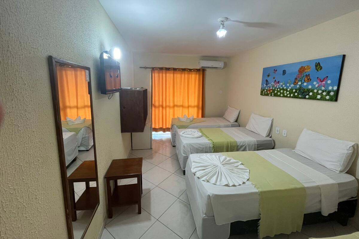 Quarto de hotel com duas camas, uma mesinha, ventoinha no teto, aparelho de ar condicionado e um quadro na parede.