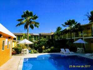 A pool area with lounge chairs and umbrellas, surrounded by palm trees and a building with a tiled roof.