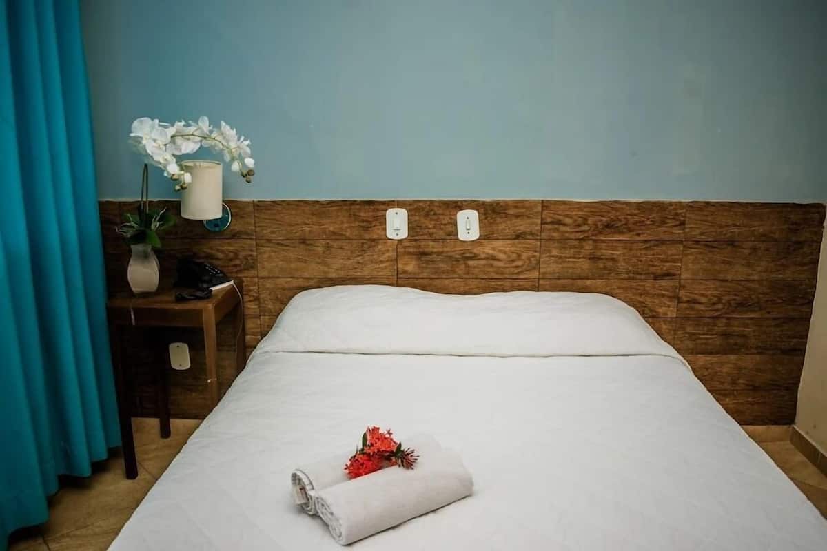 Cama bem arrumada com edredom branco, toalha enrolada e um pequeno buquê de flores vermelhas. Apresenta cabeceira de madeira, mesa de cabeceira com luminária e um vaso com flores brancas.