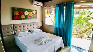 Ein Schlafzimmer mit einer gebürsteten Polsterung am Kopfteil, einem Blumenbild, einem Bett mit weißen Leinen und Blick auf Grün durch ein Fenster mit blauen Vorhängen.