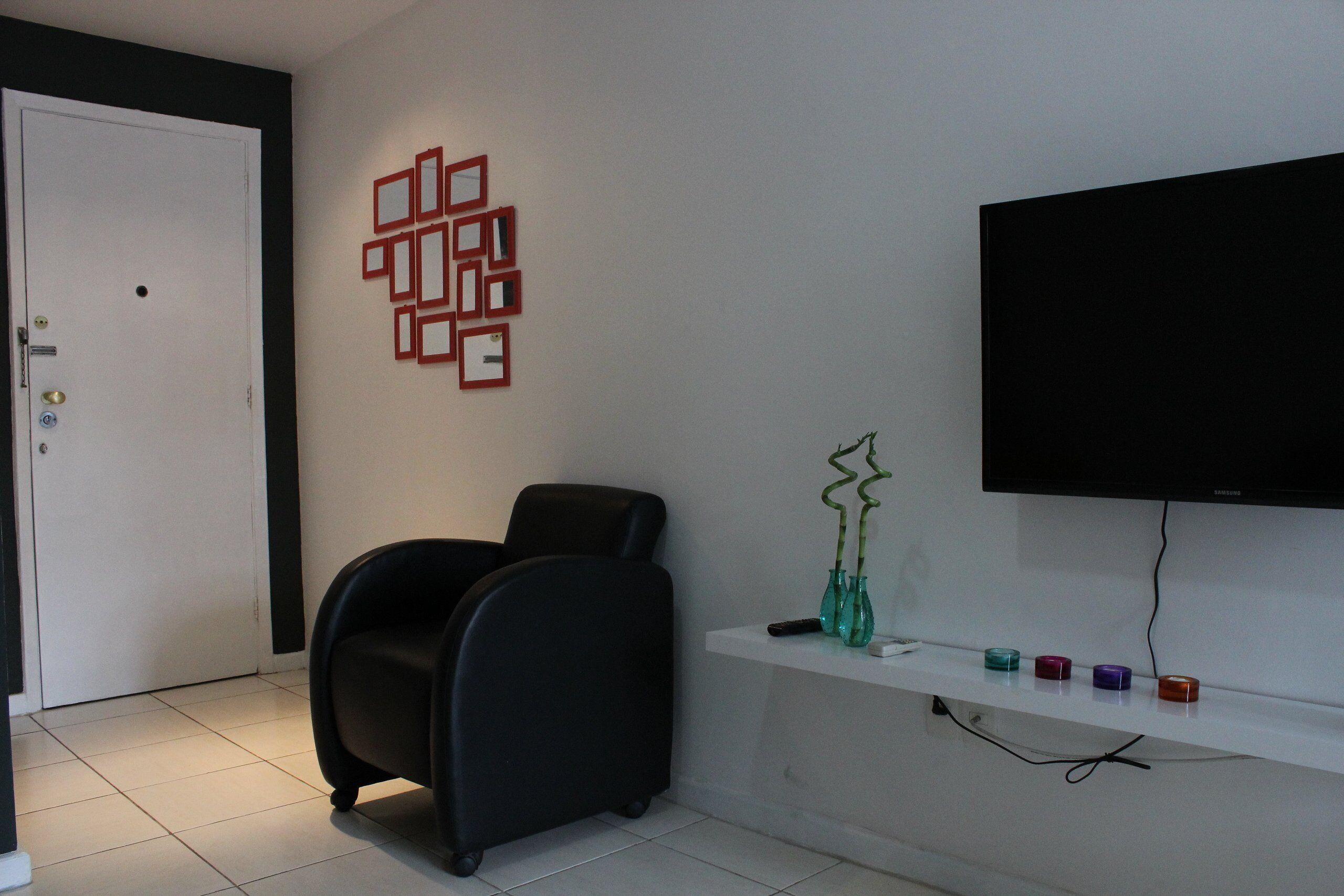 Ein Zimmer mit schwarzem Bürostuhl, einer weißen Tür, einem an der Wand montierten Fernseher und einem Regal mit Dekoration.