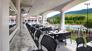 Un área de comedor al aire libre con techado, mesas y sillas, con vista a una piscina y abundante vegetación.