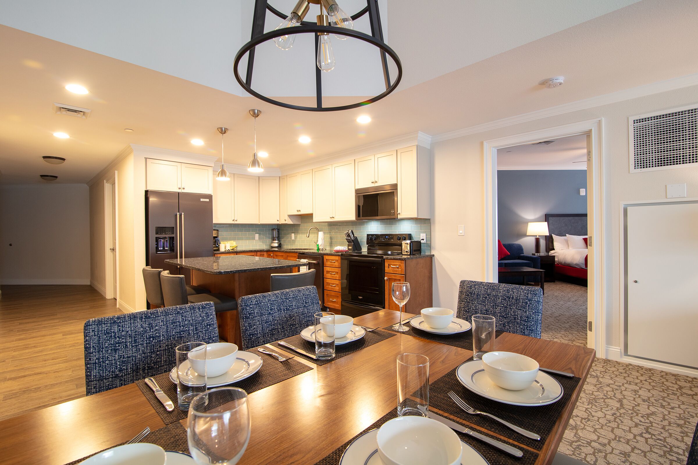 A modern kitchen with a central island, dining area with a wooden table set for four, and a living area with a sofa and lamp.