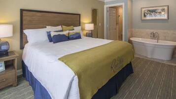 Habitación de hotel con una cama grande, una mesita de noche con lámpara, un baño con bañera y un cuadro en la pared.