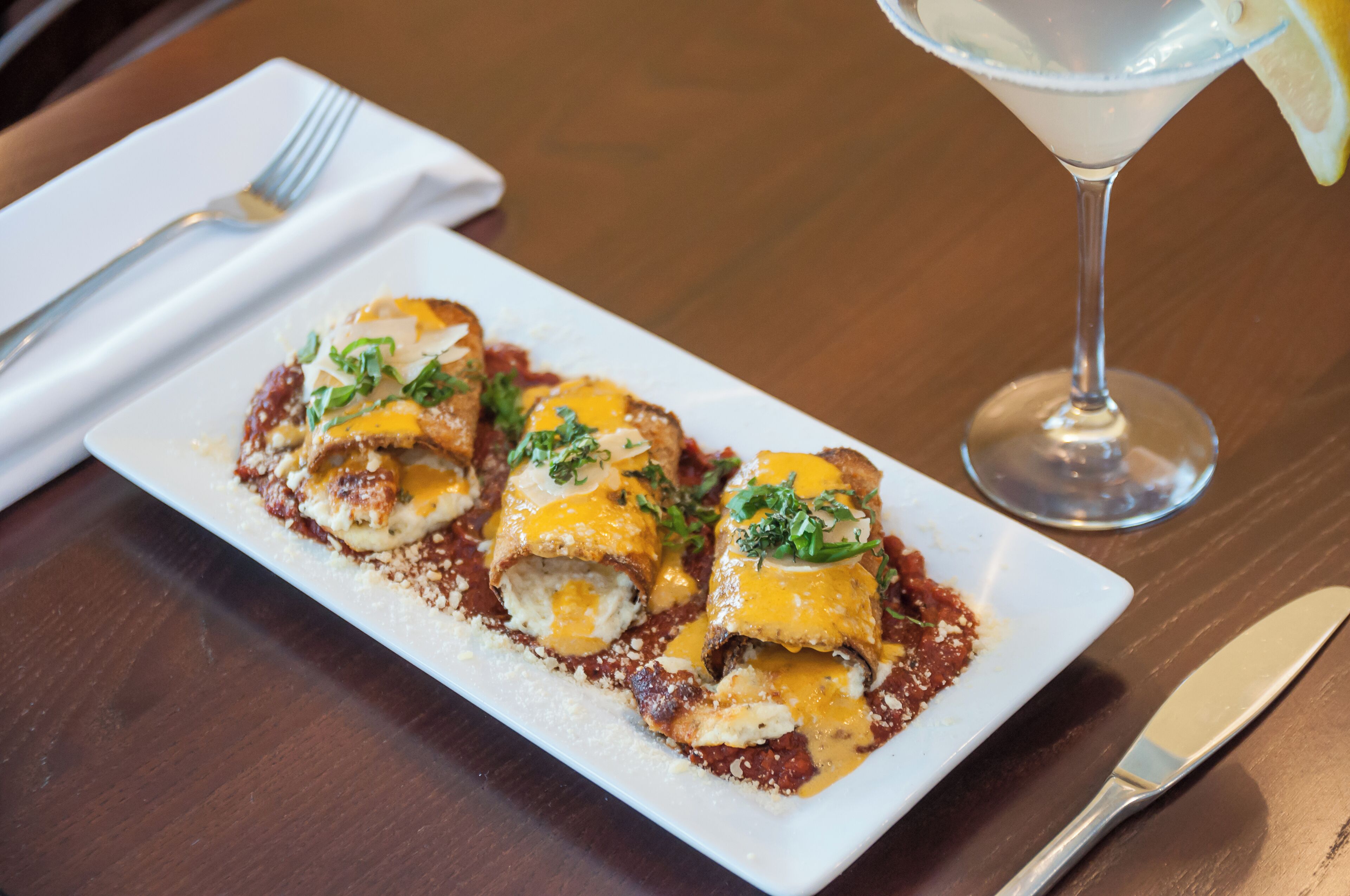 A rectangular plate with three stuffed pasta rolls, garnished with herbs, and a glass of a clear beverage with a lemon slice on the rim.