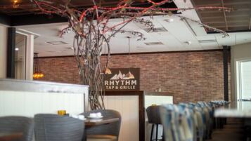 Een restaurantinterieur met een bakstenen muur, een bord met de tekst 'RHYTHM TAP & GRILLE' en rijen barkrukken.