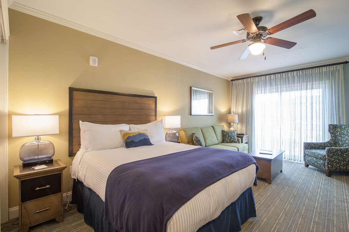A hotel room with a large bed, a ceiling fan, a nightstand with a lamp, a chair, and a window with curtains.