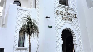 L’extérieur de l’hôtel La Coupole se distingue par sa façade blanche, ses arches décoratives et une enseigne indiquant le nom de l’hôtel.