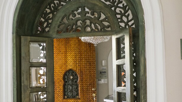 Un portail en arc surmonté de motifs métalliques complexes et un sol à carreaux menant à une pièce dont les murs sont recouverts d’un papier peint à motifs et qui est ornée d’un lustre.