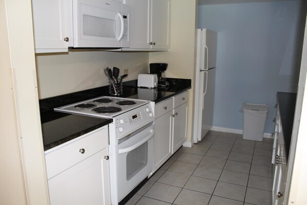 A compact kitchen with white cabinets, a black countertop, a stove, a microwave, a coffee maker, and a refrigerator.