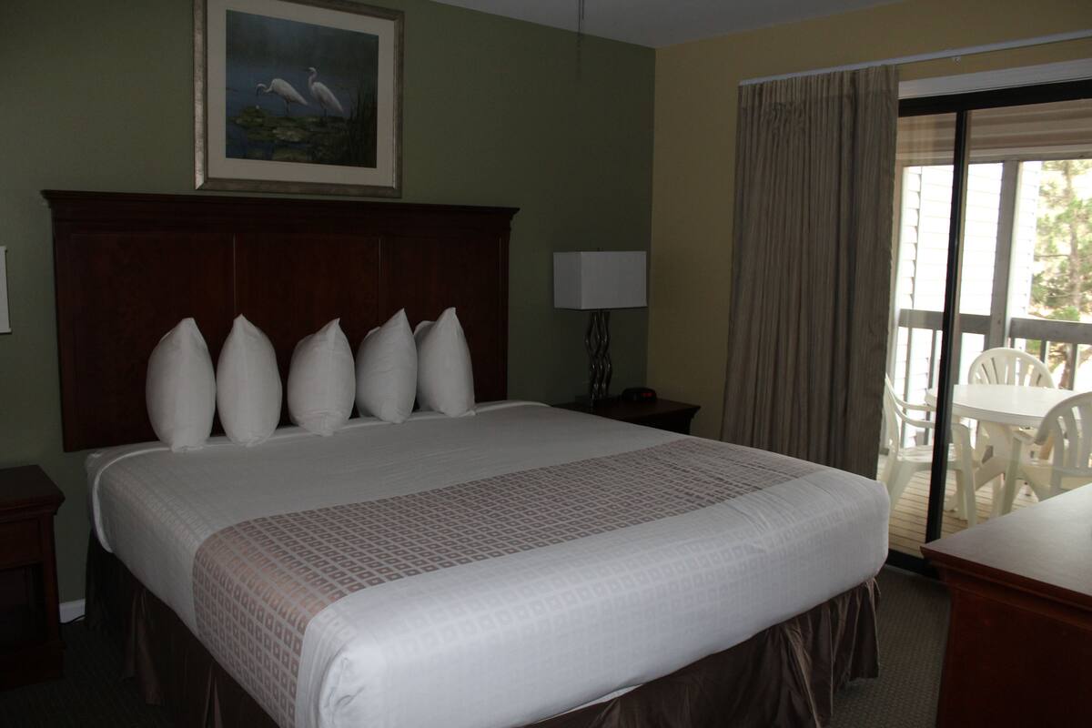 A hotel room with a large bed, a wooden headboard, a painting above the bed, a lamp on a bedside table, and a view of the outdoors through a sliding door.