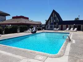 Una piscina rectangular con agua azul cristalina, rodeada por una terraza de hormigón. Hay sillas de descanso y una mesa en la terraza.