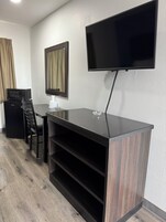 Una habitación moderna con televisión de pantalla plana en la pared, un escritorio de madera oscura con superficie de vidrio, una silla de oficina negra, un microondas y un refrigerador pequeño.