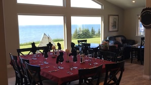A dining area with a table set for six, large windows offering a view of the ocean, and a ceiling fan. - Alaska SeaScape Lodge (Kenai)
