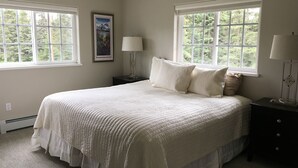 A bedroom with a large bed, two bedside tables, a nightstand, a framed picture, and two windows with views of greenery. - Alaska SeaScape Lodge (Kenai)