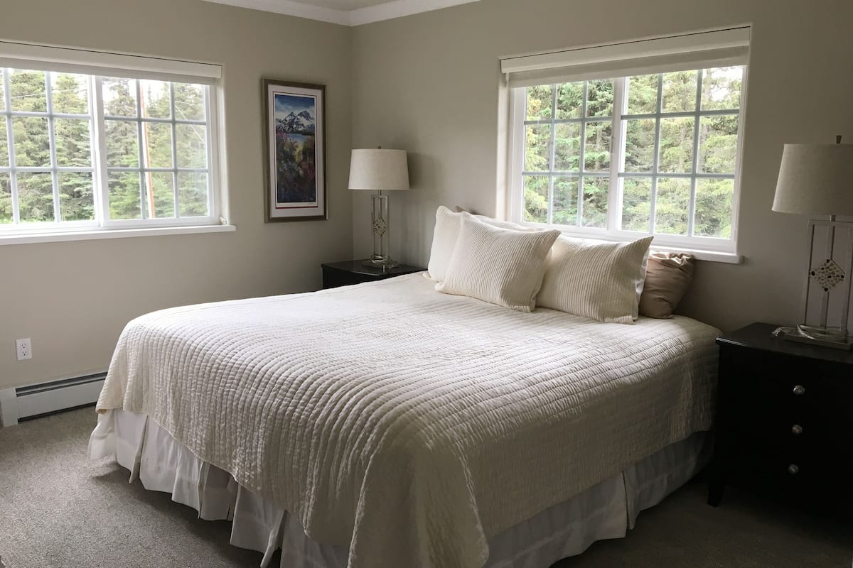 Une chambre à coucher avec un grand lit, deux tables de chevet, une commode, un tableau encadré et deux fenêtres donnant sur un paysage verdoyant.