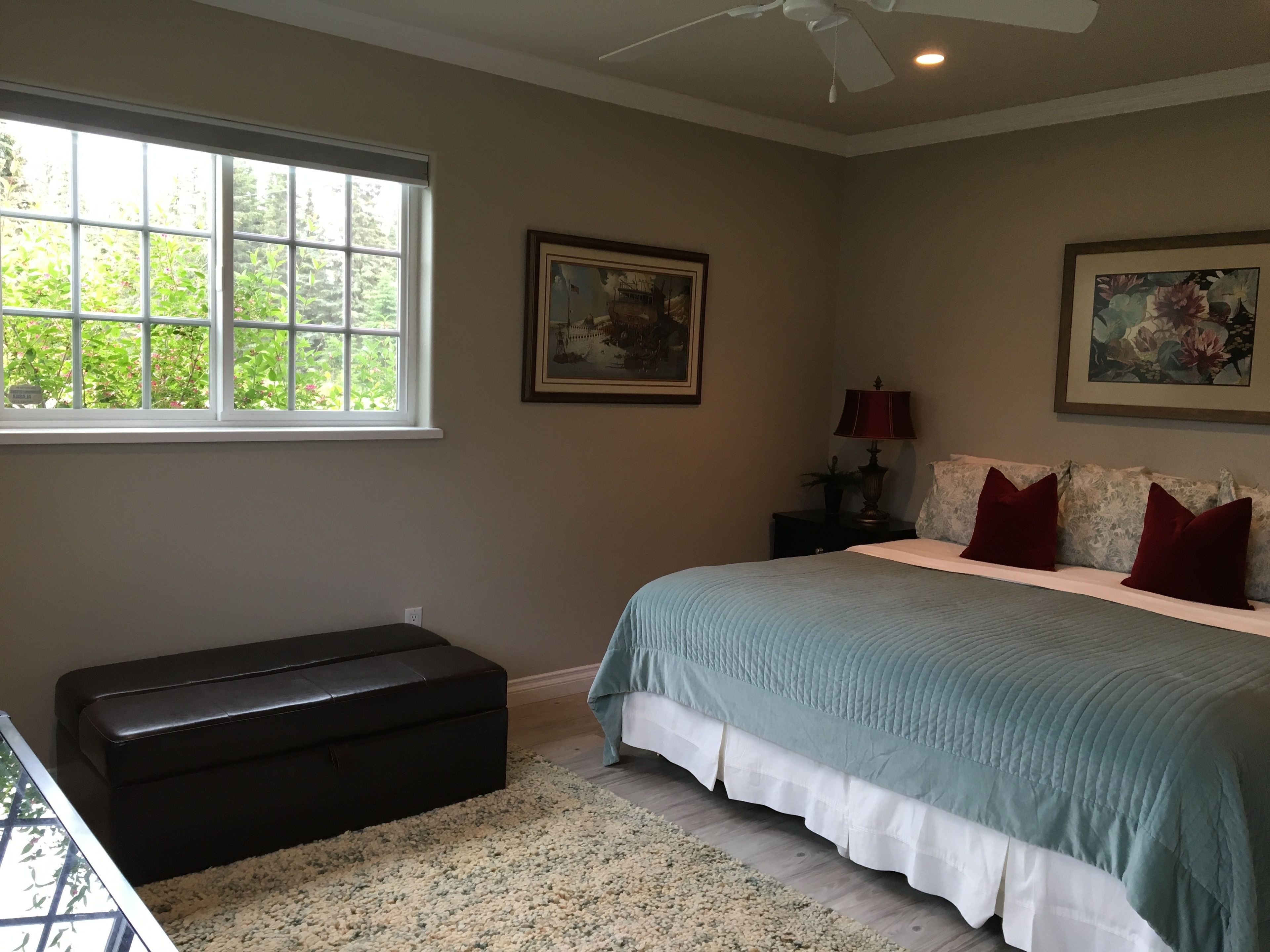 A bedroom with a large bed, a nightstand, a lamp, a framed picture, and a window with a view of greenery.