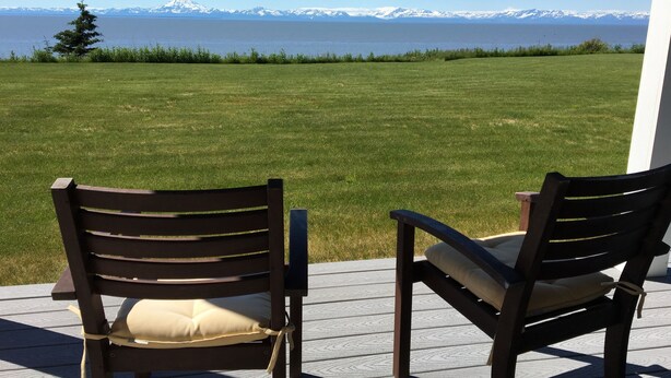 Two wooden chairs with cushions on a wooden deck overlooking a grassy area with a view of snow-capped mountains across a body of water. - Alaska SeaScape Lodge (Kenai)