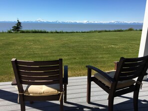 Two wooden chairs with cushions on a wooden deck overlooking a grassy area with a view of snow-capped mountains across a body of water. - Alaska SeaScape Lodge (Kenai)