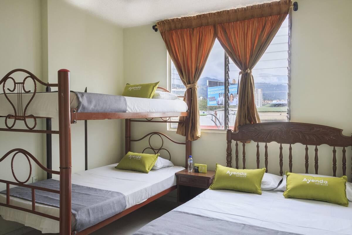 A hotel room with bunk beds, two green pillows with "Ayenda Hotels" branding, a window with curtains, and a view of a cityscape.