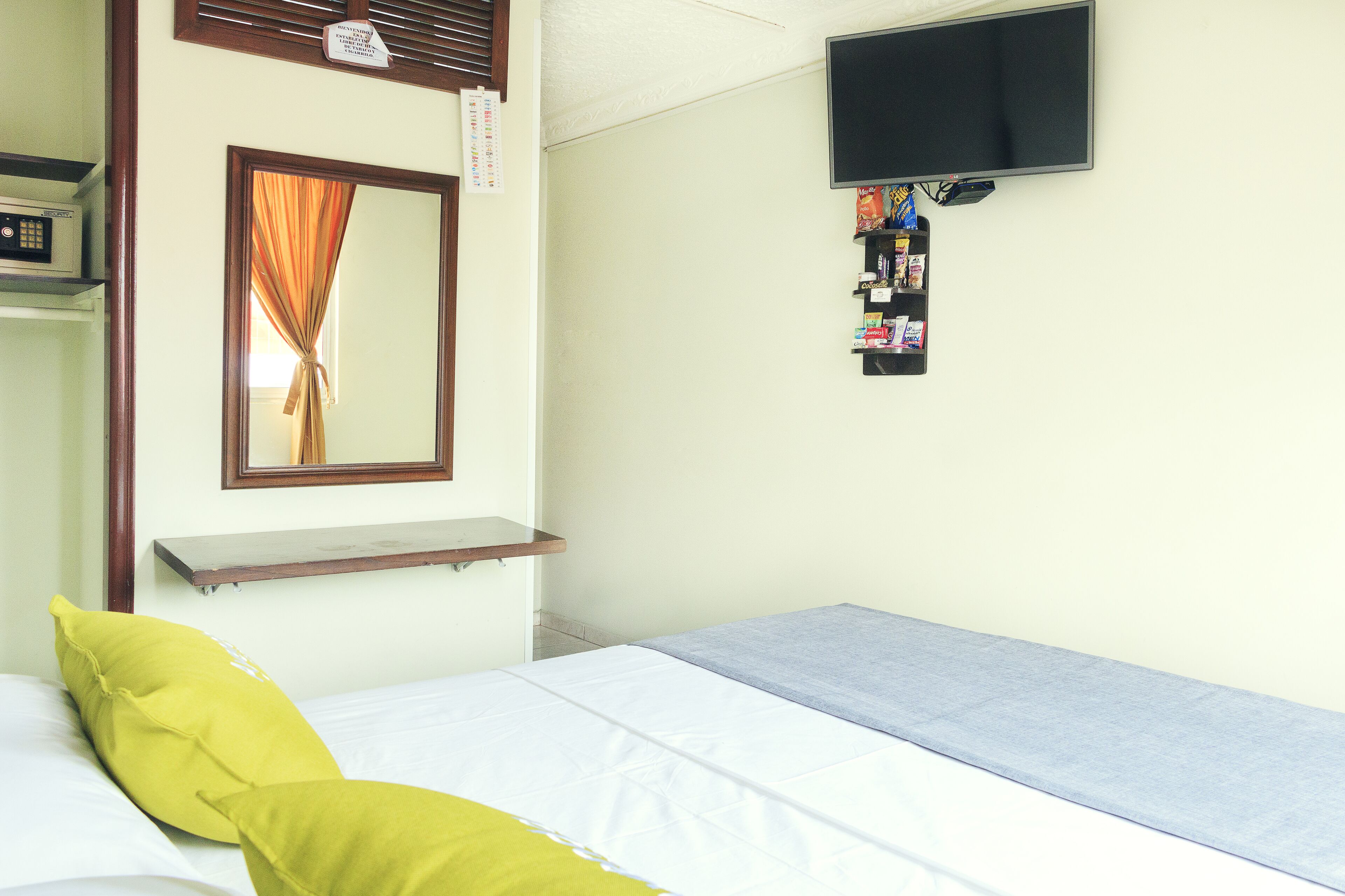 Una habitación con una cama, un televisor en la pared, una estantería con objetos y una ventana con cortinas.