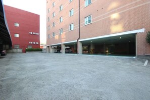 A parking area with multiple parking spaces and a covered parking structure adjacent to a multi-story building with a red facade. - Utopia Motel (Gumi)