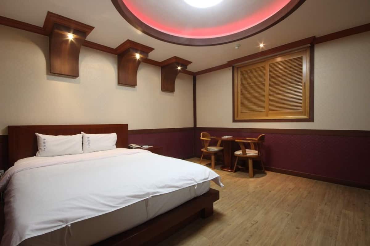 A hotel room with a large bed, a desk, two chairs, a window with blinds, and a ceiling light.