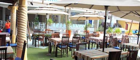 Un área de comedor al aire libre con varias mesas y sillas, cubierta por sombrillas y rodeada de plantas en macetas.