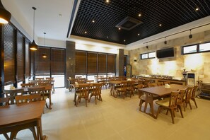 A spacious dining area with multiple tables and chairs, large windows with blinds, and a kitchen area with appliances.