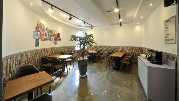 A modern restaurant with a polished floor, wooden tables, and chairs. There is a large potted plant in the center, and a menu board on the right wall.