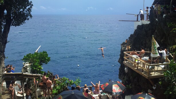 Escena costera con un acantilado sobre el mar, una terraza de madera con gente y vistas al océano.