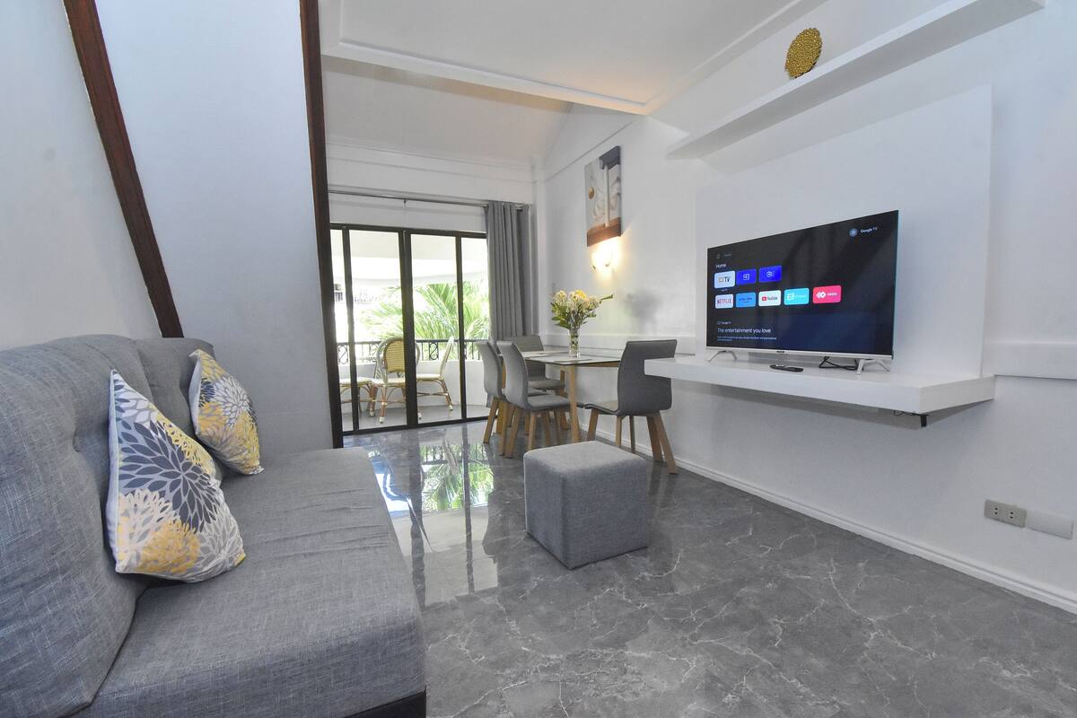 Un salón moderno con un sofá gris, un reposapiés y un televisor de pantalla plana montado en una pared blanca.