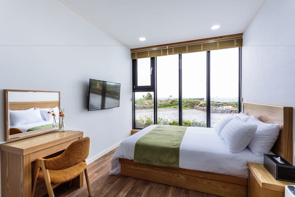 Une chambre d’hôtel avec un grand lit, un bureau en bois, une chaise, un téléviseur et une vue sur l’extérieur grâce à de grandes fenêtres.