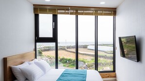 Een slaapkamer met een groot raam, een bed met wit beddengoed, een houten hoofdbord en een flatscreen televisie aan de muur.