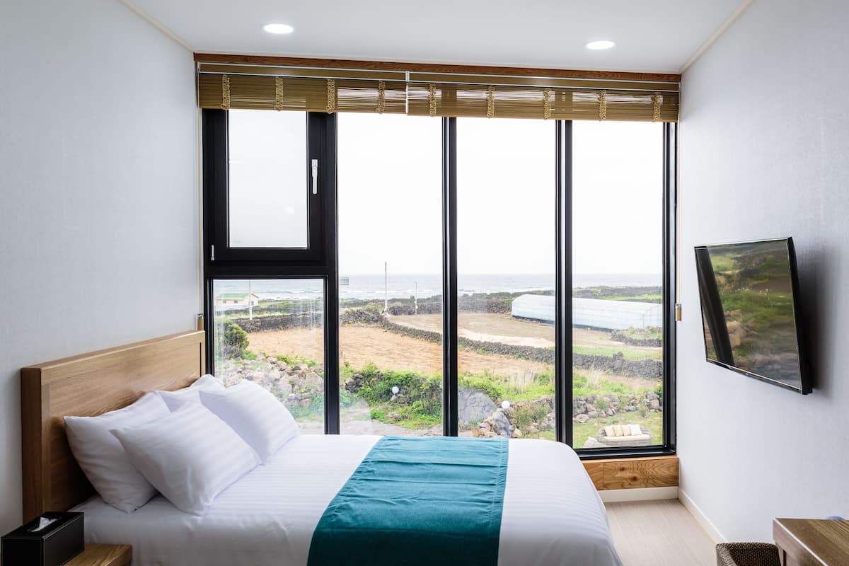Une chambre dotée d’une grande fenêtre, d’un lit avec une literie blanche, d’une tête de lit en bois et d’un téléviseur à écran plat fixé au mur.