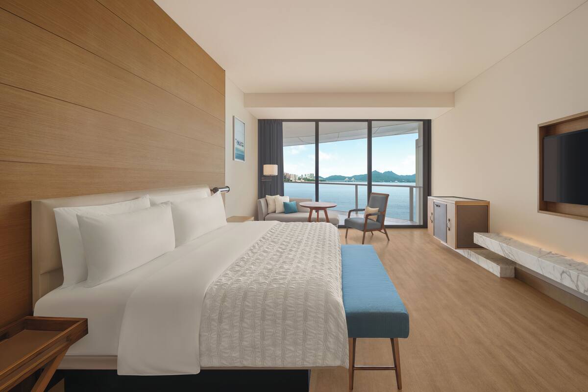 Ein modernes Hotelzimmer mit einem großen Bett, einer Sitzecke mit Tisch und Stühlen, einem an der Wand befestigten Fernseher und Blick auf das Wasser.
