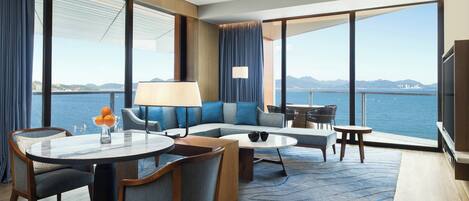 A modern hotel room with a large balcony, a sofa, a dining table, and a TV.