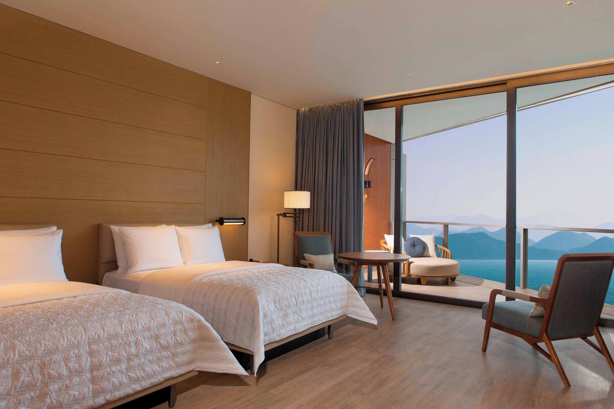 Ein Hotelzimmer mit zwei Betten, einer Sitzecke mit Tisch und Stühlen sowie einem großen Fenster mit Blick auf die Berge.
