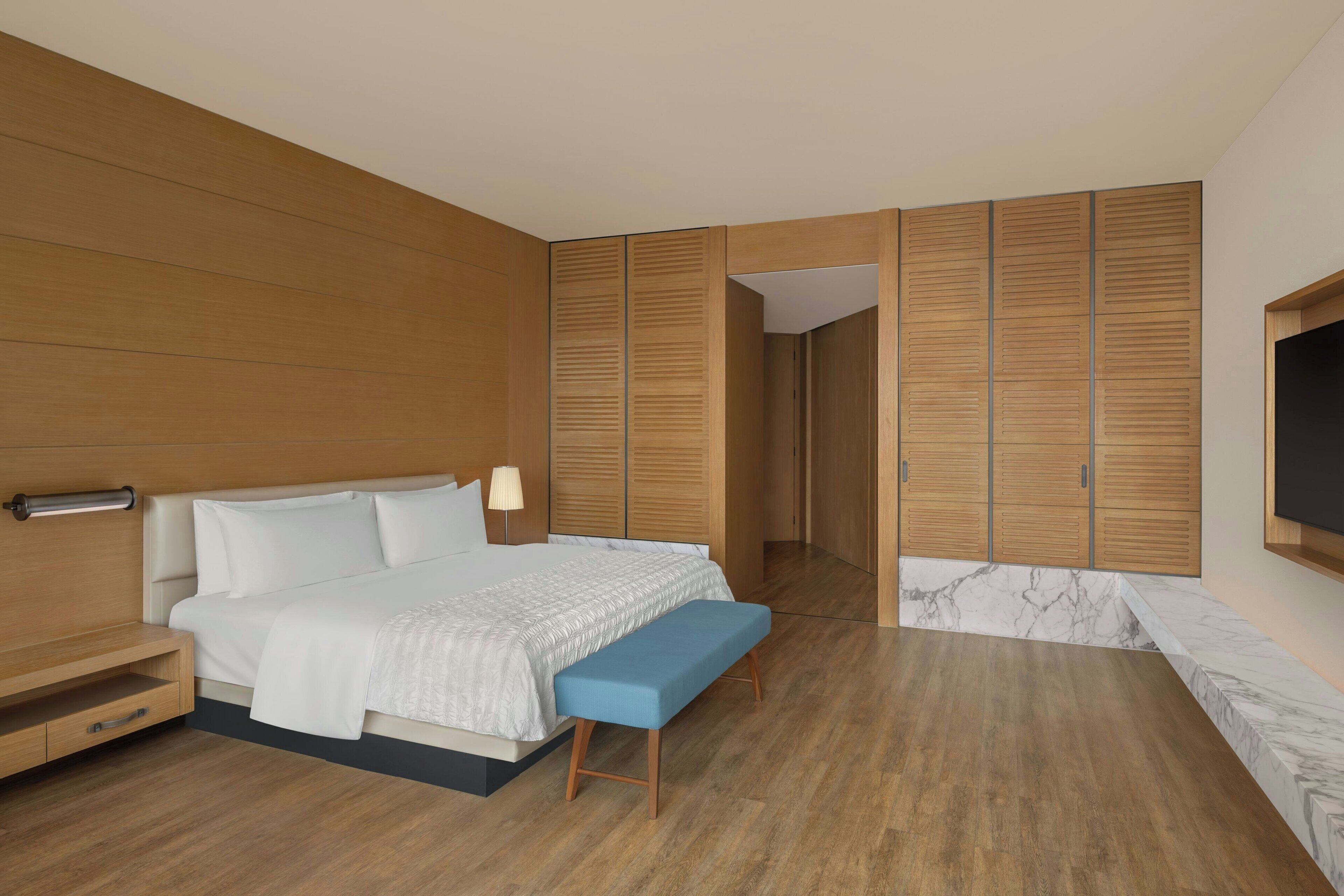 Un dormitorio moderno con cabecera de madera, cama con ropa de cama blanca, un banco azul, armario de madera y una pared de acento de mármol.