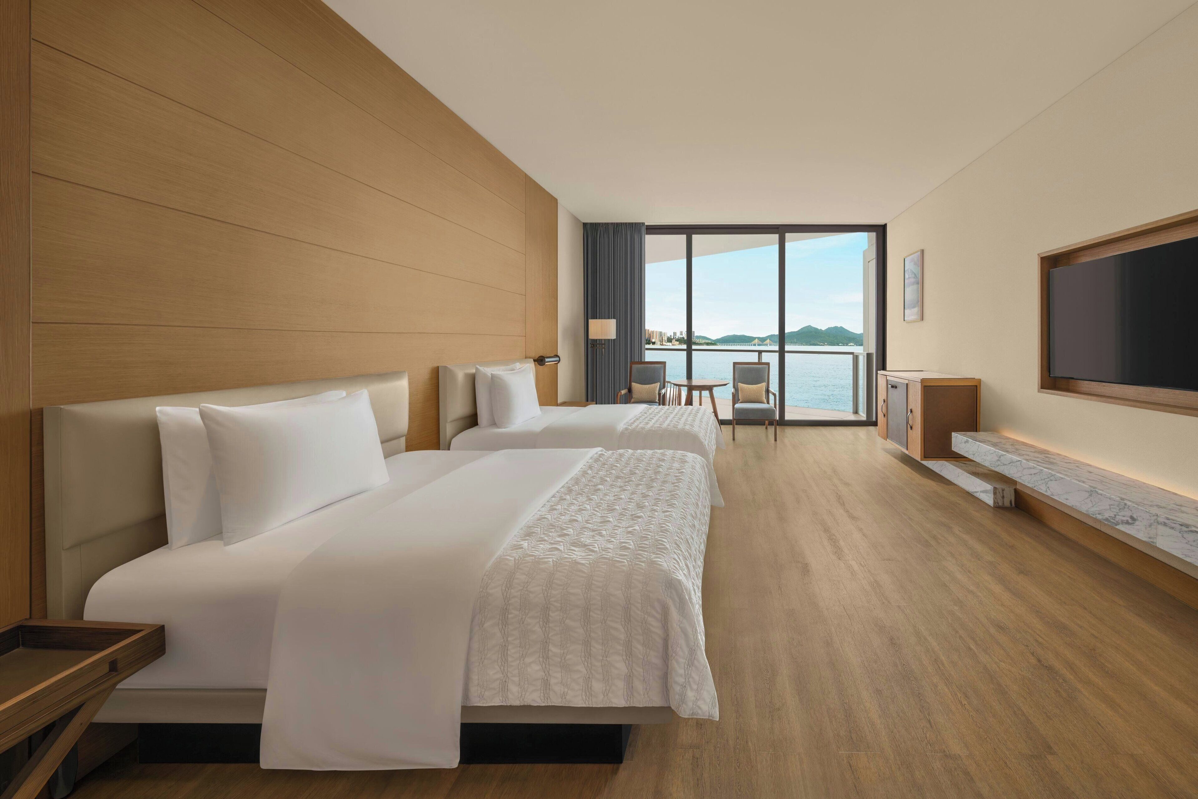 A hotel room with two beds, a TV, a balcony with a view of the sea, and a wooden floor.