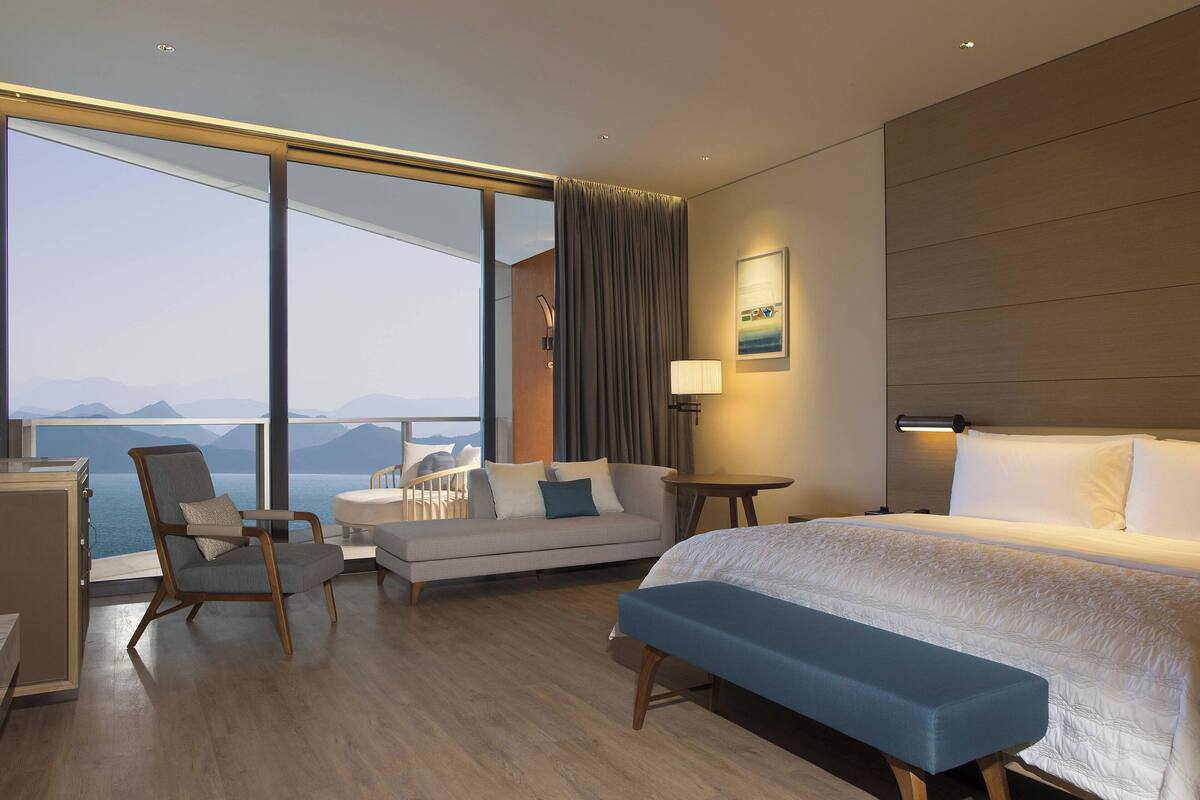 Ein modernes Hotelzimmer mit großem Fenster, einem Bett, einer Couch, einem Sessel und einem Schreibtisch.