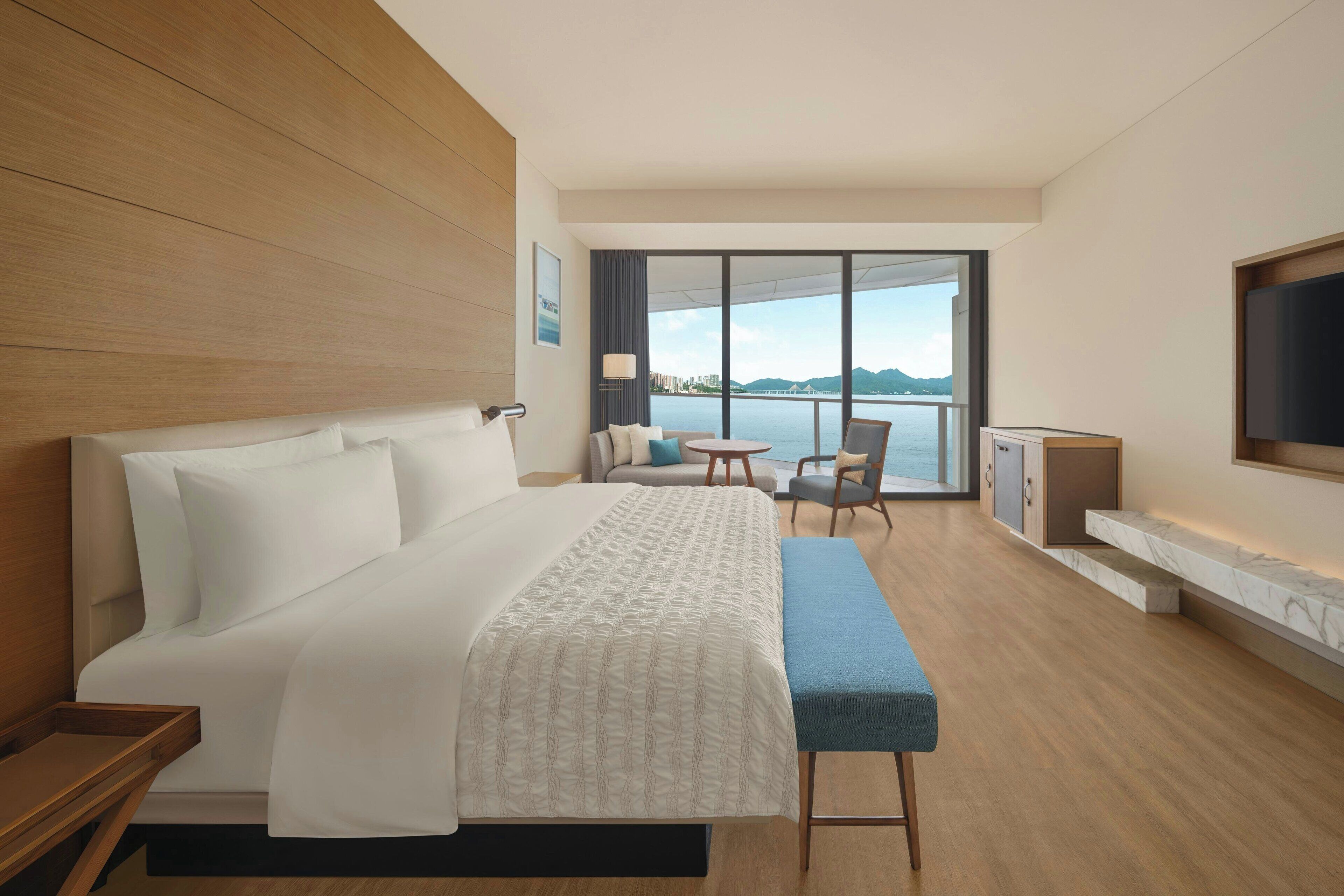 Quarto de hotel moderno com uma cama grande, área de estar com mesa e cadeiras, TV fixada na parede e vista para o mar através de grandes janelas.